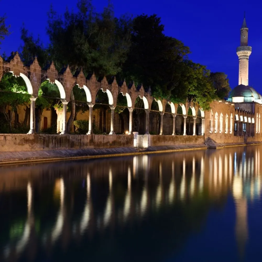 Balıklıgöl in Şanlıurfa at dusk, with the lit arcades and the minaret reflected in the pool