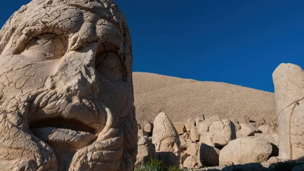Colossal stone head on the Mount Nemrut terrace, with the tumulus of Antiochus I and other statues behind under a blue sky